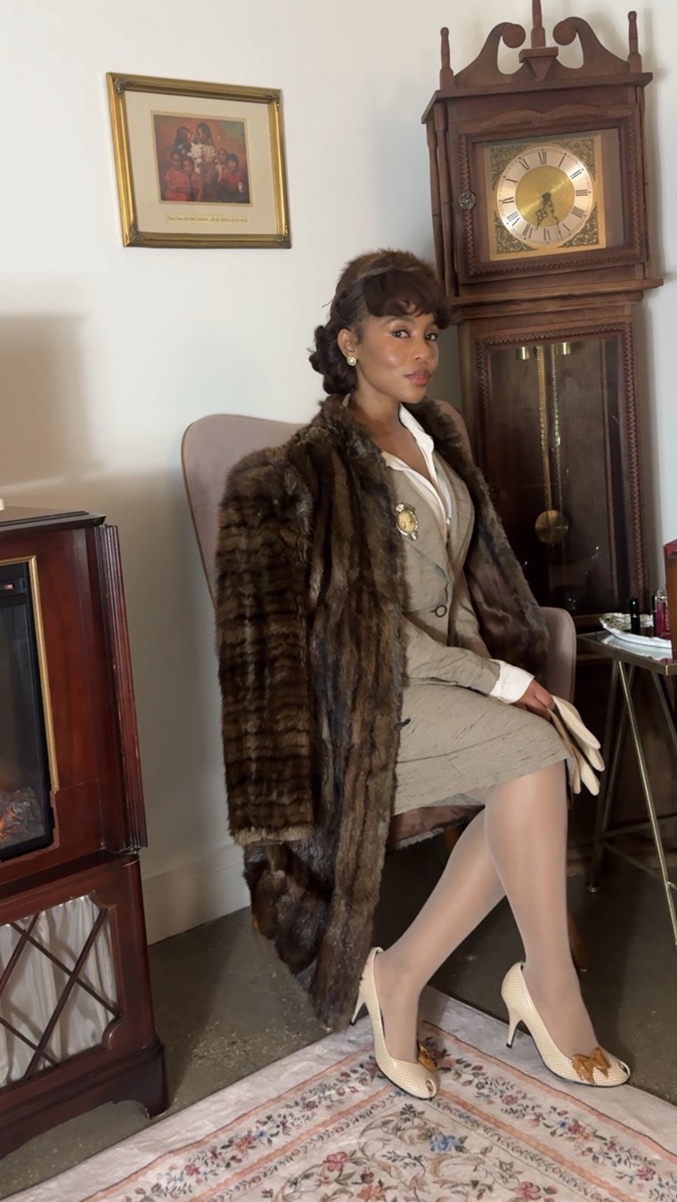 Fur-collar coat over a tweed suit, seated